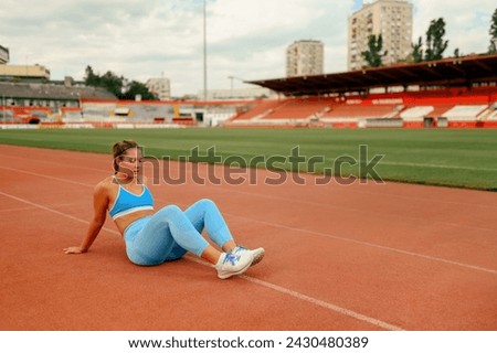 A fit sportswoman is sitting on running track at stadium and taking a break from running. A tired female runner in shape is sitting at stadium and resting. Healthy lifestyle, healthcare, bodycare.