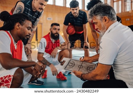 A senior basketball coach is holding clipboard with game tactics while his multicultural team is listening to him and watching game plan on training. Coach is working on game plan with basketball team Royalty-Free Stock Photo #2430482249