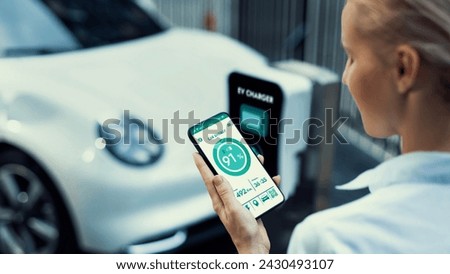Businesswoman holding smartphone display battery status interface by smart EV mobile application while EV car recharging electricity from charging station in car park. Peruse