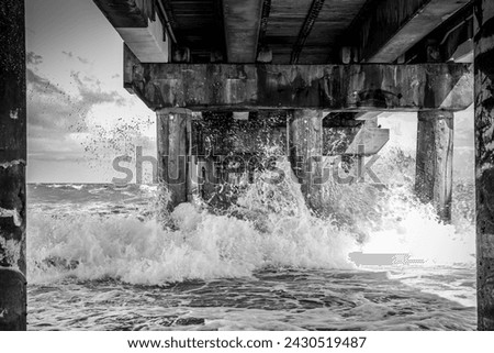 Dynamic monochrome photograph showcasing the power of the ocean as waves crash under a pier, ideal for editorial use in nature magazines, environmental campaigns, and as dramatic wall art.