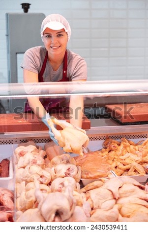 Smiling skilled young female butcher working behind counter in butchery, showing fresh raw farm chickens Royalty-Free Stock Photo #2430539411