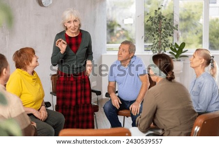 Older woman presides over support group meeting with mature people listening Royalty-Free Stock Photo #2430539557