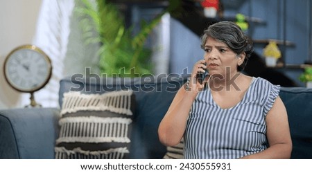 Upset Indian old grey-haired woman sitting on sofa couch talking on mobile phone answer unpleasant call received bad shocking news. Disappointed worried senior grandmother feel sad alone indoor home Royalty-Free Stock Photo #2430555931