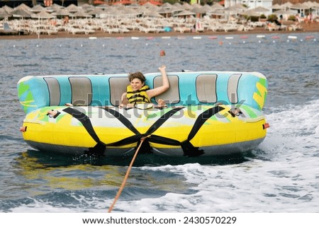 Young boy exudes joy while riding an inflatable tube towed by a boat in the ocean. Happy school child having fun in adventure water park on the sea. Royalty-Free Stock Photo #2430570229
