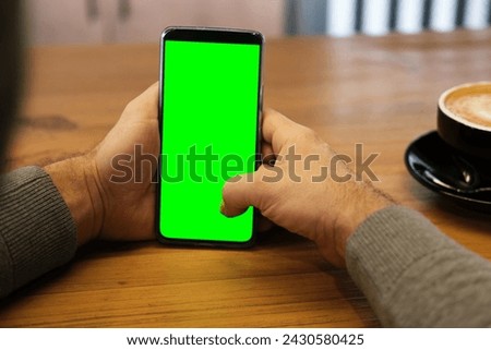 Young man sitting at cafe holding smartphone green mock-up screen in hand. Male person using chroma key mobile phone. Vertical mode. Touching, swiping display, tapping, surfing internet social media