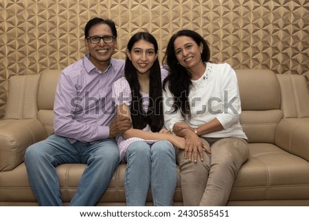 Indian happy mature couple and young girl are hugging, looking at camera and smiling while sitting on couch at home. Concept of Happy Daughter Day.