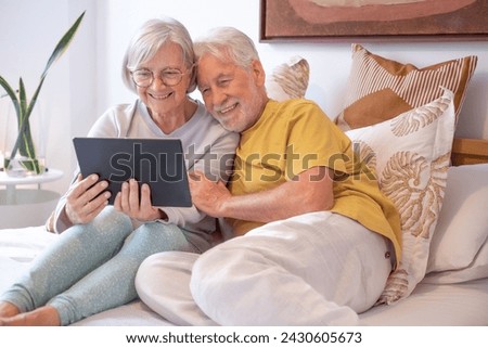 Video call concept. Portrait of happy senior couple communicating in remote chat with family and friends - Grandparents with digital tablet sitting in bed, wireless technology