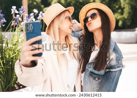 Two young beautiful smiling hipster female in trendy summer clothes. Carefree women posing in the street. Positive models having fun outdoors at sunny day. Cheerful and happy. In hat, take selfie