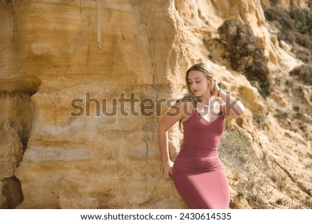 Young, beautiful, blonde woman in a pink dress, sitting on a stone wall, hand in her hair, looking at the ground relaxed and calm. Concept innocence, tenderness, virginity.