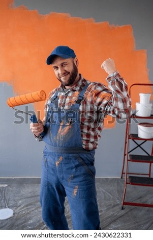 Adult bearded painter man in blue overalls over orange wall background celebrating a victory in winner position. Copy space