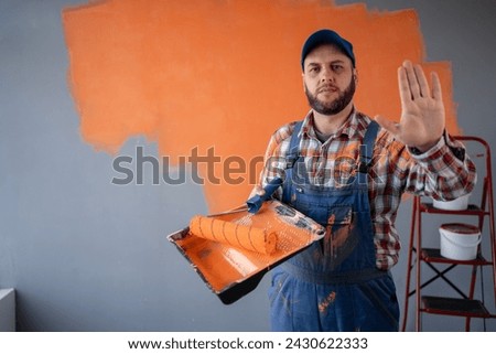 Painter man holding a paint roller and tray making stop gesture. orange wall background