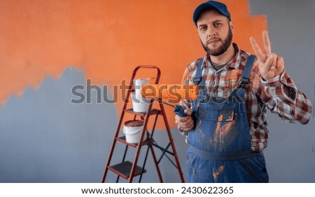 Adult bearded painter man in blue overalls over orange wall background celebrating win showing sign of victory. Copy space