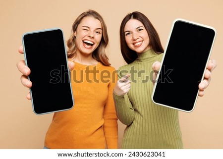Young friend two women they wear orange green shirt casual clothes together hold use close up mobile cell phone with blank screen workspace area isolated on plain beige background. Lifestyle concept
