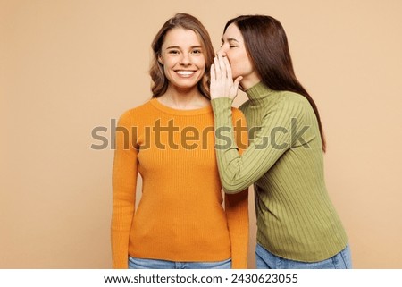 Young friends two women they wear orange green shirt casual clothes together whispering gossip and tell secret behind her hand sharing news isolated on plain pastel beige background. Lifestyle concept