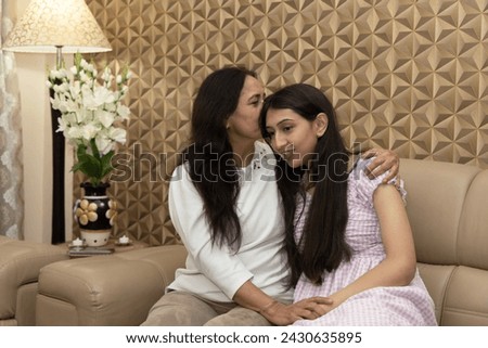 Happy Indian mid-aged woman kissing daughter with closed eyes in forehead and holding hands with young female while sitting on couch at home together