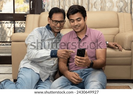 Happy Indian mid-aged father and young son using on phone while sitting on sofa at home. Concept of social media , lifestyles and internet connection. smartphone booking or buying online on mobile