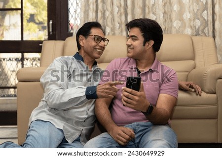 Happy Indian mid-aged father and young son using on phone while sitting on sofa at home. Concept of social media , lifestyles and internet connection. smartphone booking or buying online on mobile