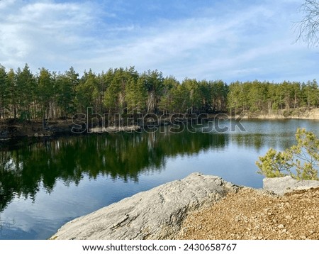 A clear transparent lake with granite rocks reflects a blue sky with white clouds and green trees