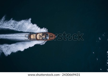 Expensive wooden boat, man and woman in motion on the water making a white trail looking like air. A large modern high-speed wooden luxury boat moves on blue water, top view. Royalty-Free Stock Photo #2430669431