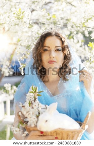 A beautiful girl in a blue dress with a decorative white rabbit against the background of a blooming apple tree. Animals and people.