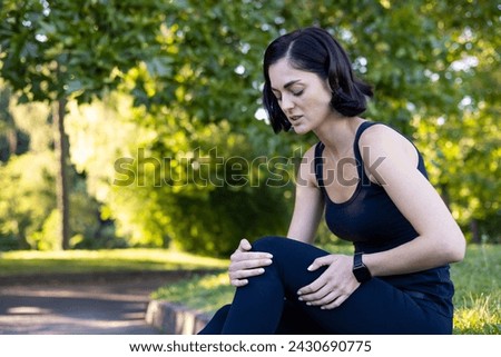 Photo of a young woman doing sports and jogging in the park, sitting on the curb and holding her leg with her hands. Feels severe knee pain and sprain.