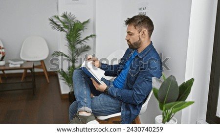 Relaxed young hispanic male settling into a waiting room chair, engrossed in a book - a handsome portrait of rest, focus, and ongoing pursuit of knowledge. indoor corridor forms a serene background. Royalty-Free Stock Photo #2430740259