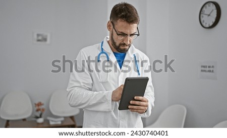Handsome young hispanic man, a dedicated doctor, immersed in work on his touchpad, sitting relaxed yet serious in a clinic waiting room, epitome of professional healthcare.