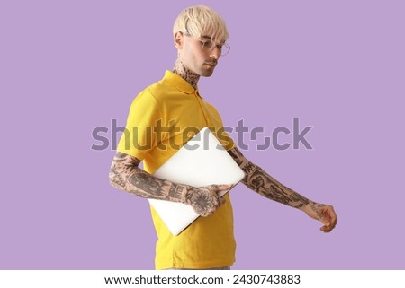 Tattooed young man with laptop on lilac background