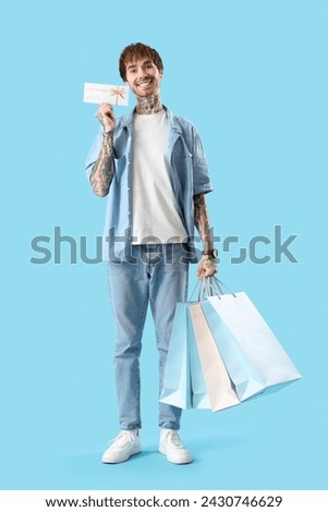 Young tattooed man with gift card and shopping bags on blue background