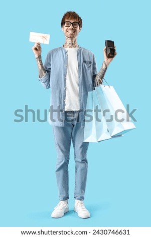 Young tattooed man with gift card, payment terminal and shopping bags on blue background