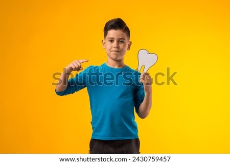 Portrait of child boy holding papercraft tooth and pointing finger at it over yellow background. Dental health concept
