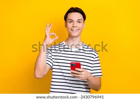 Photo of attractive guy dressed striped t-shirt hold smartphone showing okey approve good quality isolated on yellow color background