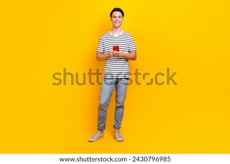 Full size photo of intelligent student dressed striped t-shirt jeans trousers holding smartphone isolated on yellow color background