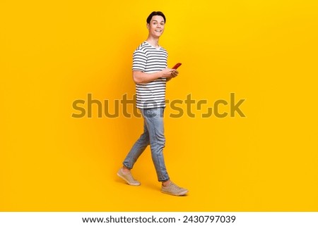 Full size photo of optimistic student dressed striped t-shirt hold smartphone go to empty space isolated on yellow color background