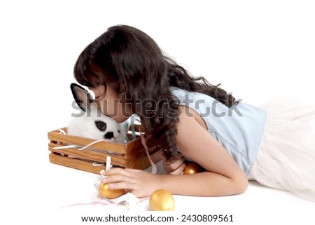 Cute little girl kissing rabbit in wooden box. Joyful kid lies down on white background with golden easter eggs and her pet, celebrating Easter day. Happy child on spring holiday celebration.