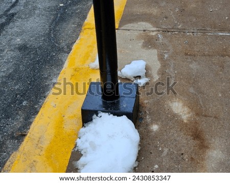 The base of a black pole along a sidewalk with snow around it.