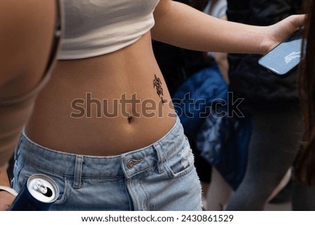 Close-up of a woman's midriff with a henna tattoo, holding a can, with a smartphone and a blurred background