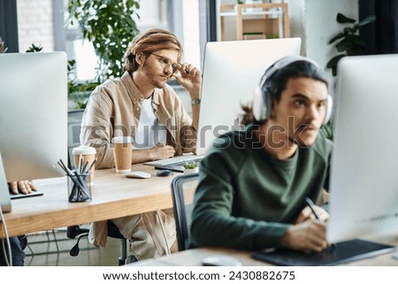 pensive male professional adjusting glasses and looking at monitor near blurred coworker