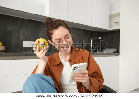 Young woman chilling at home, looking at smartphone, reading news on social media and eating green apple in the kitchen.