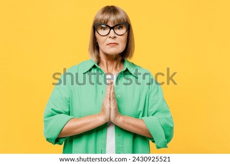 Elderly blonde woman 50s years old wear green shirt glasses casual clothes hold hands folded in prayer gesture, begging about something isolated on plain yellow background studio. Lifestyle concept Royalty-Free Stock Photo #2430925521