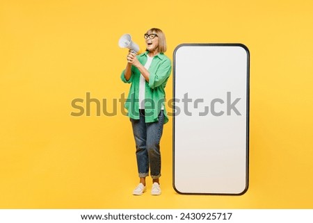 Full body elderly blonde woman 50s year old wear green shirt glasses casual clothes big huge blank screen mobile cell phone smartphone with area scream in megaphone isolated on plain yellow background