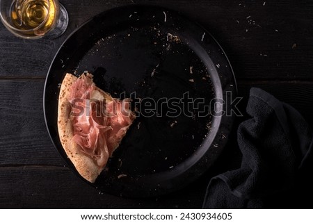 Slice of pizza with prosciutto and mozzarella cheese on a black plate with a glass of white wine. View from above.