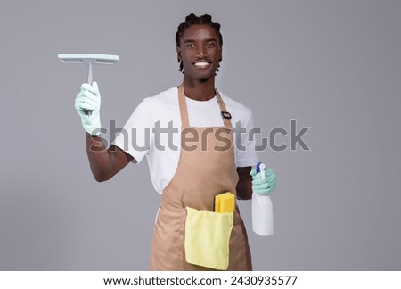 Smiling Black Cleaner Man In Apron Advertising Cleaning Services, Handsome African American Guy With Detergents And Washing Tools In Hands Standing Against Grey Studio Background, Copy Space
