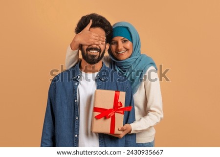 Loving muslim woman in hijab covering eyes and surprising her husband with gift box over beige studio background, greeting him with birthday, wedding anniversary or Valentine's day, copy space