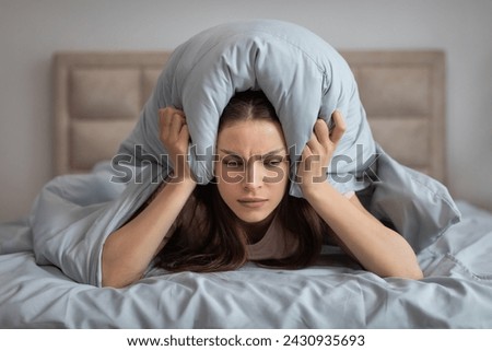 Troubled young woman lying in bed clutches her pillow over her head, her expression one of frustration and annoyance, as she attempts to block out the disruptive noise interrupting her rest Royalty-Free Stock Photo #2430935693