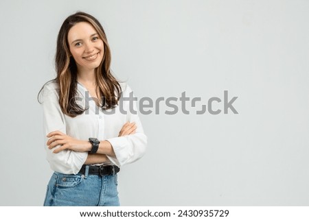 A confident young european woman with long brunette hair smiles gently at the camera, arms crossed, dressed in a white blouse and blue jeans, with a smartwatch on her wrist Royalty-Free Stock Photo #2430935729