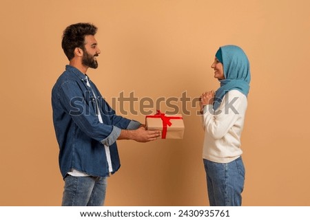 Loving arab man giving wrapped gift box to his delighted muslim girlfriend, romantic millennial husband greeting wife in hijab with birthday or valentines day, standing against beige background