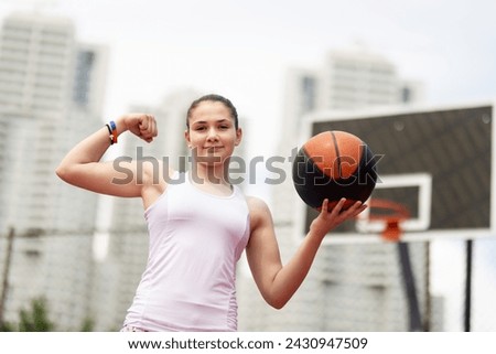 Basketball player portrait. Girl posing and showing muscles. Outdoor sports field with urban background.