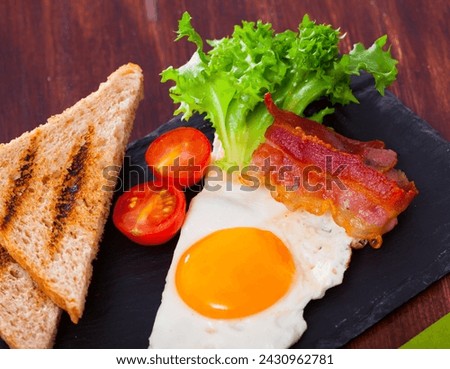 Picture of fried eggs with bacon, toasted bread, tomatoes and lettuce at plate on table
