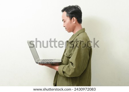 portrait of asian man holding and looking at laptop screen with smile face. Indonesian man in green shirt on isolated background. illustration of a businessman, entrepreneur, freelancer or student